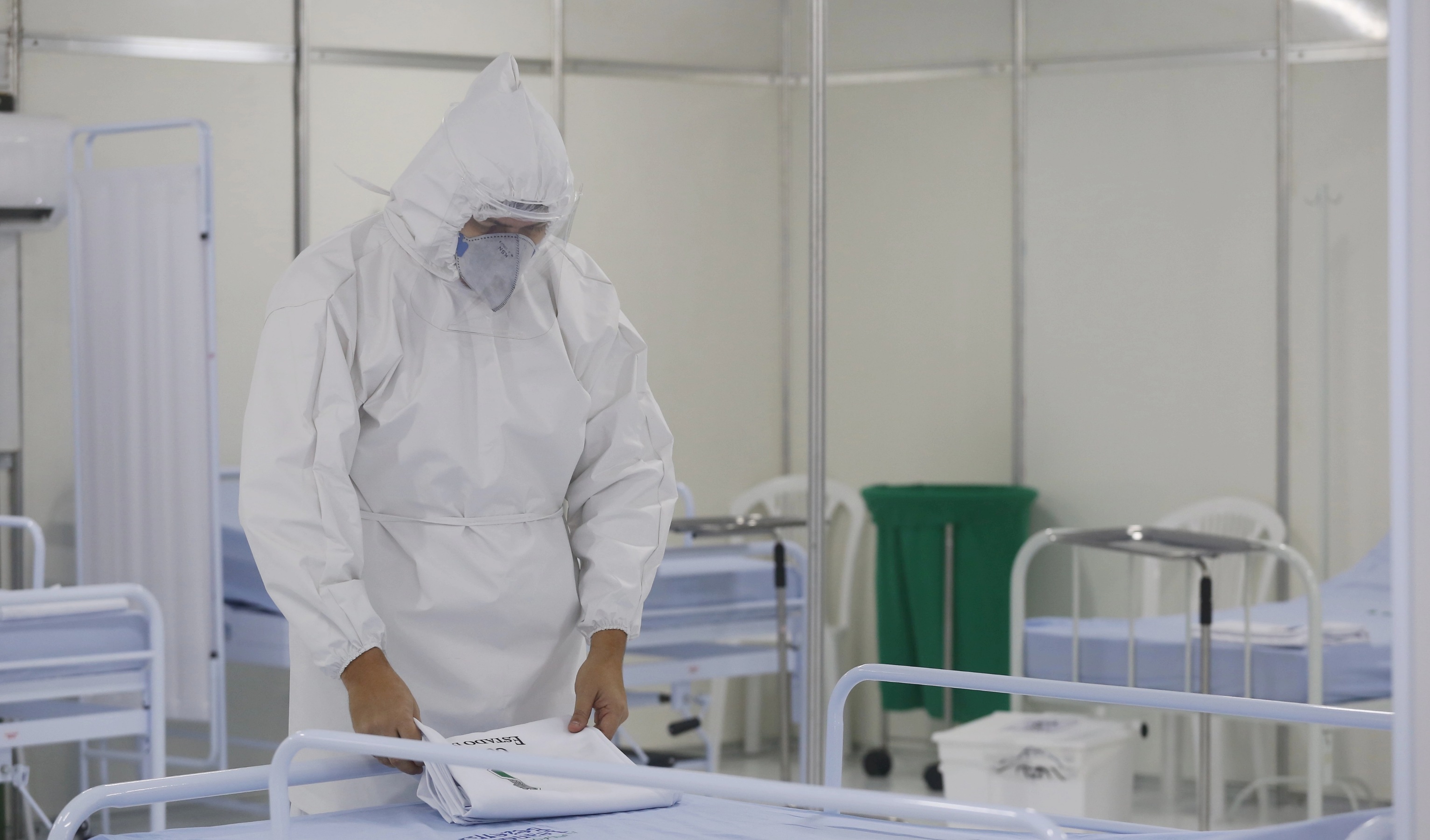 homem usando roupa de médico, máscaras, touca dobrando lençol em cima de leito de hospital